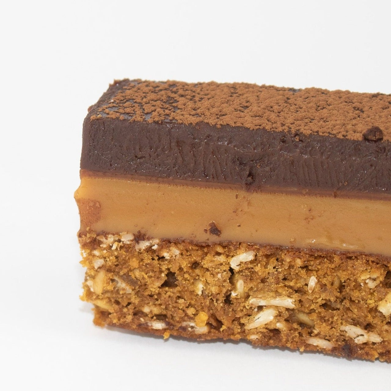 Close up image of a 3 layer slice. the bottom layer is an oaty biscuit. the middle layer is caramel. and at the top there is a thick layer of chocolate ganache