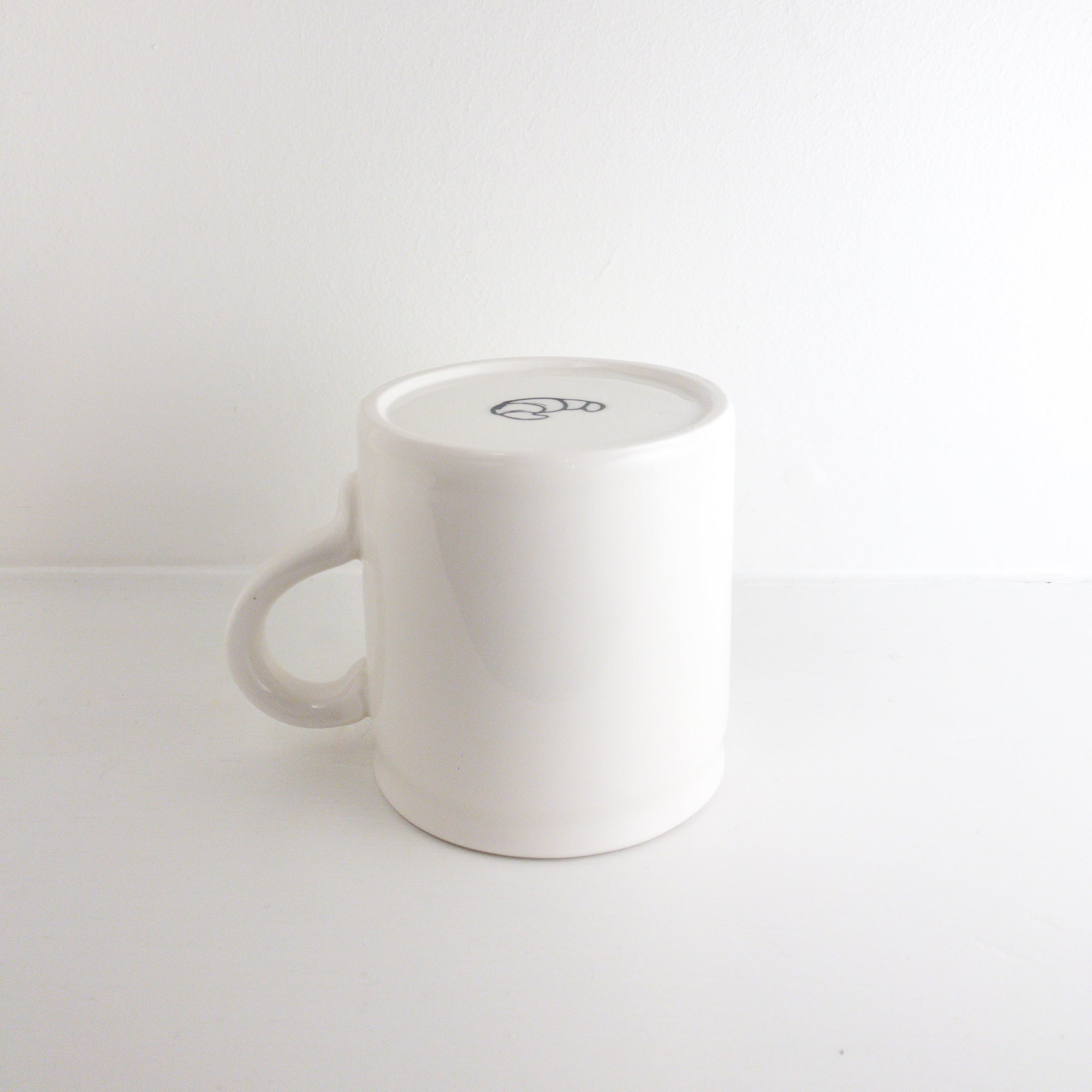 image of a handmade white mug upside down. it shows a small croissant on the bottom 